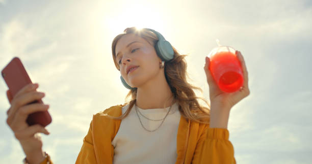 musica, telefono e donna in estate con succo e cuffie ascoltando la web radio. cielo blu, streaming audio mobile e podcast online con una giovane persona della gen z che si sente rilassata all'aperto durante una vacanza - tourist resort audio foto e immagini stock