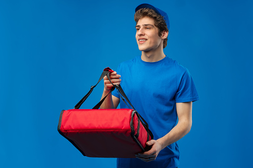 Young courier with thermo bag on blue background in studio