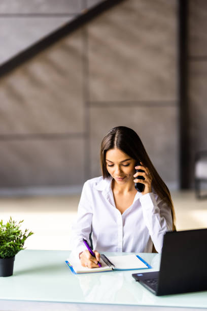 istock Attractive smiling young businesswoman wearing jacket talking on mobile phone while sitting on a desk and using laptop in office 1475472549