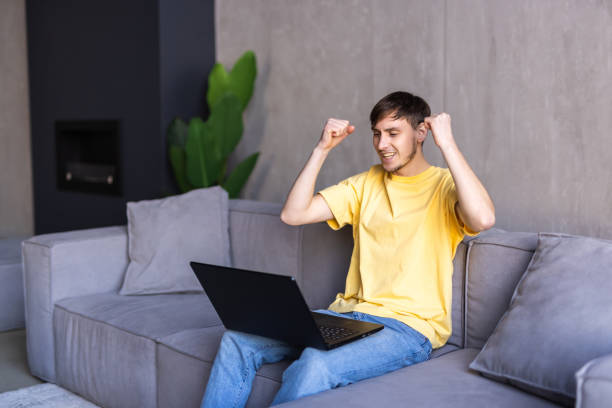 istock Excited young man with laptop at home sitting on a couch 1475478100