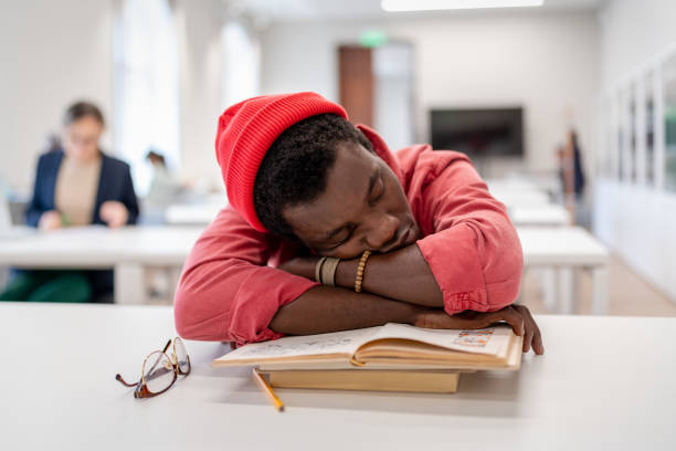 African male student falling asleep during study in library, sitting at desk sleeping on books African male student falling asleep during study, feeling drowsy while studying in library, selective focus. Black guy sitting at desk sleeping on textbooks, napping in classroom. College burnout male college student sleeping through lecture stock pictures, royalty-free photos & images