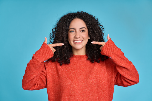 Happy young latin woman pointing at white healthy teeth, showing perfect dental toothy smile isolated on blue background advertising dentistry tooth care whitening clinic services concept.