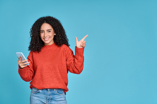 Young happy latin woman holding mobile phone pointing aside isolated on blue.