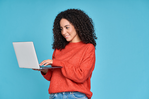 Young latin woman student using laptop device standing isolated on blue background. Smiling female model user holding computer, typing, surfing, searching job online or shopping website.