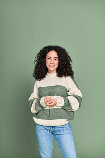 Young adult stylish beautiful smiling latin woman, happy pretty female model student wearing striped sweater and jeans standing looking at camera isolated on green background, vertical portrait.