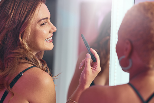 Makeup, girl friends and getting ready by a mirror with a smile ready for a party. Lipstick, cosmetics and happy person laughing with cosmetic product and skin glow smiling with a friend at a club