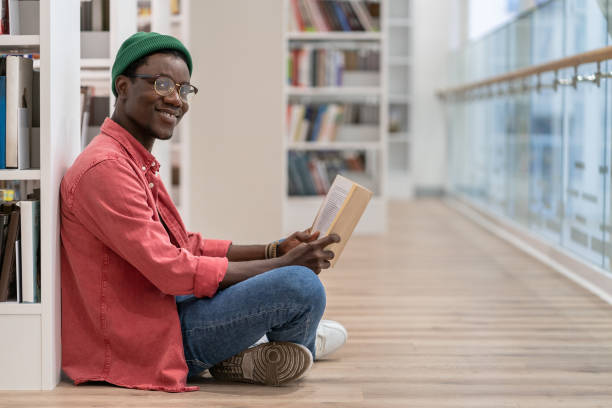 istock Smiling Black student man reading book in university library , looking at camera. Reading hobby. 1475484513