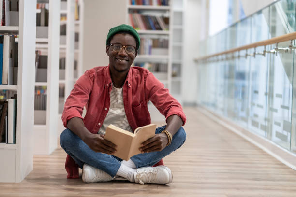 istock Happy clever black student man with book sitting on floor in university library, looking at camera 1475484521