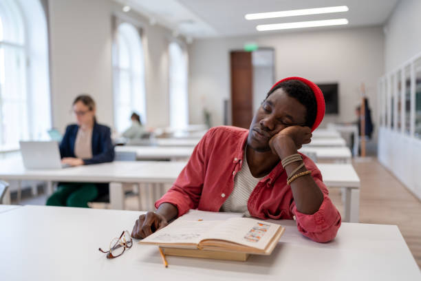 Sleepy African American man falls asleep sits in library with head resting on desk with books Sleepy African man falls asleep sits in public library tired after long time reading literature. Exhausted black guy dozing with head resting on desktop with books on university campus male college student sleeping through lecture stock pictures, royalty-free photos & images