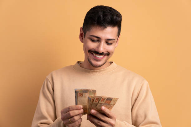 homem brasileiro alegre com notas de moeda do brasil em fundo bege. conceito de negócios, empréstimo, pagamento, riqueza. - real people - fotografias e filmes do acervo