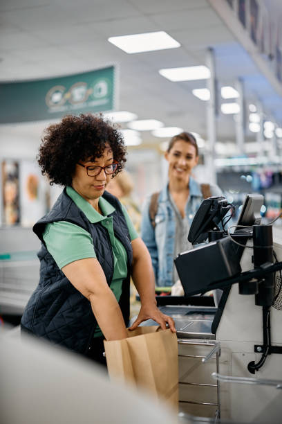 istock Supermarket worker assisting her customer with using paper bag at self-service checkout. 1475488689