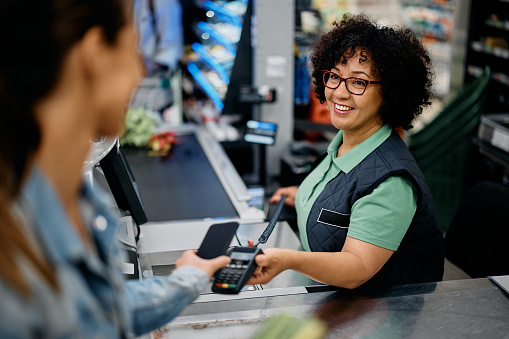 Happy cashier holding credit card reader while customer is paying with smart phone at supermarket.