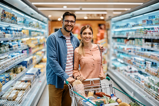 Portrait of happy couple with shopping cart full of groceries in supermarket looking at camera.