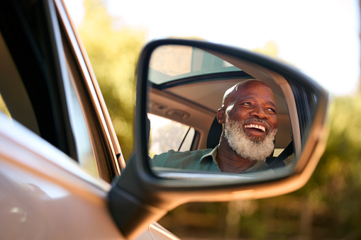 Smiling Senior Male Driver Reflected In Wing Mirror Of Car Enjoying Day Trip Out Driving
