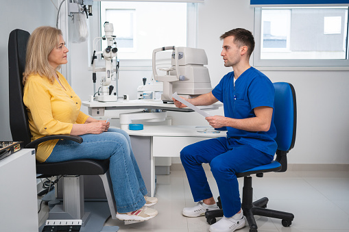 At the ophthalmology clinic, a Caucasian male ophthalmologist holding test results, while talking with a senior female patient after an exam
