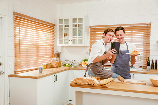 Happy Caucasian gay couple cooking together in the kitchen while watching how to cook on a digital tablet with fun and a smile. Man standing by boyfriend preparing food at home. LGBT couple concept.