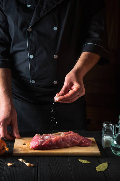 istock Chef sprinkles the raw beef meat with salt. Preparing meat before baking. Working environment in kitchen of restaurant or cafe 1475512601