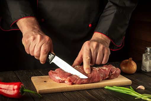 A cook cuts raw veal on a cutting board with a knife before cooking. Asian cuisine. Hotel Recipe Idea.