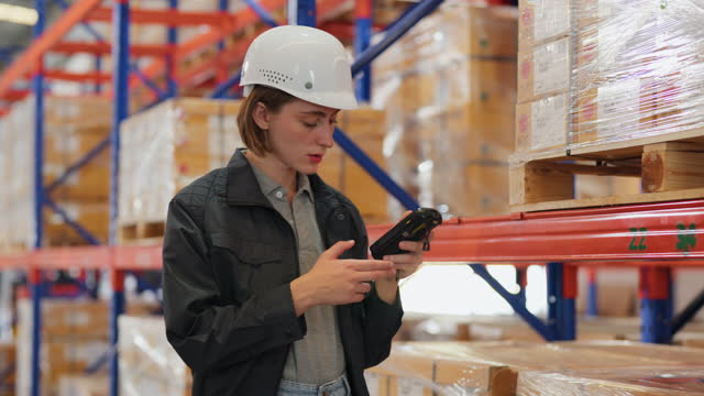 Implementing RFID tags help make delivery easier and growing up your business sustainably. Caucasian warehouse worker with safety helmet using a barcode reader to scan QR code on the box standing in the distribution warehouse. Looking at tablet for checki