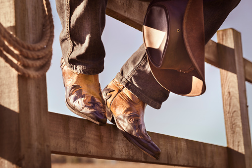 Cowboy boots and hat sitting on fence at ranch rodeo with feet up resting, country music festival live concert or line dancing concept