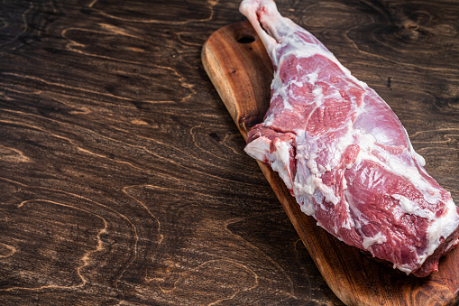 Mutton meat. Raw whole lamb leg thigh on butcher board. Wooden background. Top view. Copy space.