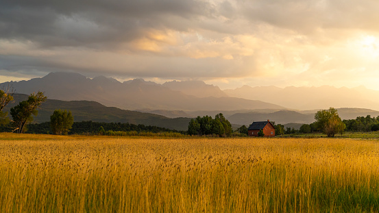 The Sun shines through the clouds after an afternoon rain in the San Juan Mountains, southwestern Colorado. Sunlight falls on the landscape below, lighting up a golden wheat field and a distant barn.