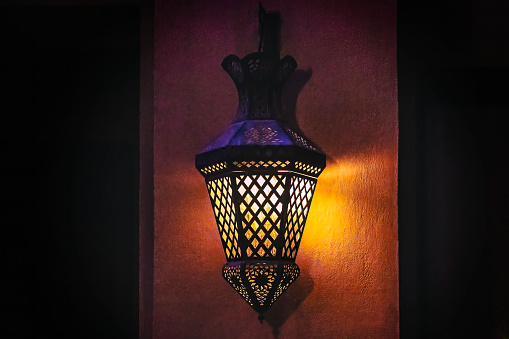 The photo depicts a modern traditional lantern on the wall which is a type of contemporary lighting fixture that combines traditional design elements with modern aesthetics. This lantern is made of high-quality metal and glass and features a sleek and streamlined design that is both functional and stylish. The yellow and orange light is very captivating. While the black space is available for copy space.