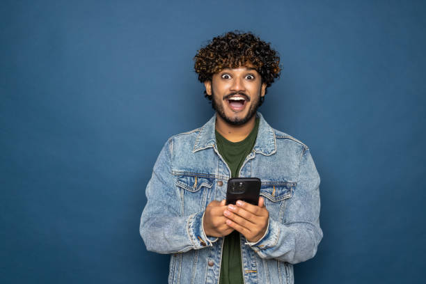 istock Young handsome Indian man using mobile phone on blue background 1475524795