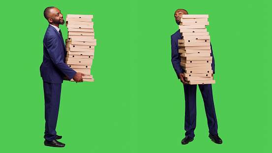 Office employee carrying big pile of pizza packs in studio, holding fast food meal over full body green screen background. Startup businessman in suit having takeout order on camera.