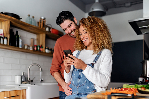 Stock photo of a middle aged woman and man standing in a kitchen and laughing. They are looking at a smartphone.