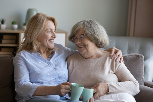 Middle-aged woman hugs her older 70s mum enjoy warm friendly talk, drinking tea smiling resting together on couch spend leisure at home. Multi-generational family bond and ties, support, relaxation