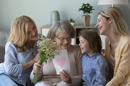 Loving mature daughter, granddaughter and great-granddaughter congratulating senior granny, make surprise on birthday or anniversary, giving flowers and postcard, express love and care. Family ties