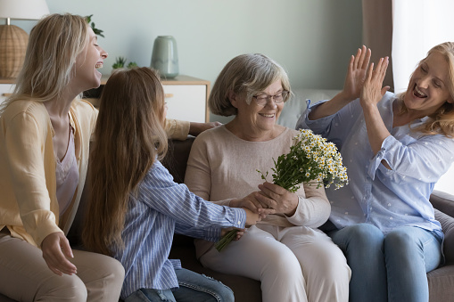 Three generations of women, little girl, adult mother and grandmother clap hands congratulating elderly great-grandmother give spring bouquet of chamomile flowers showing love and care. Family events