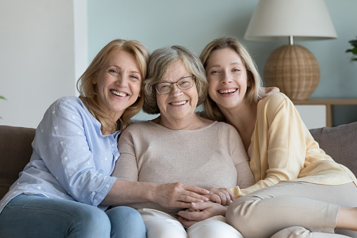 Intergenerational family portrait. Happy diverse women, elderly granny, mature daughter, millennial granddaughter smile pose for camera rest on sofa, enjoy time together, having good friendly relation