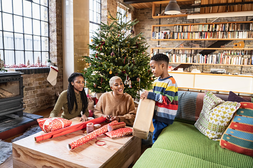 Black family together in open plan loft apartment, sitting at coffee table, smiling and learning art of gift preparation with decorated fir tree in background.