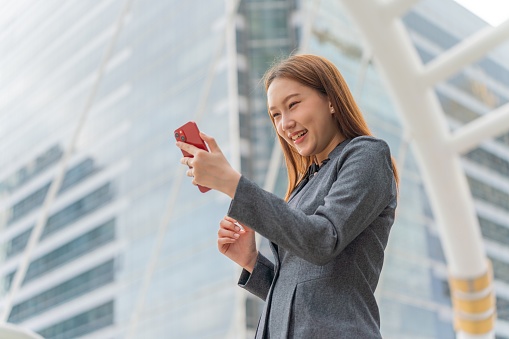 Portrait photo of a young asian businesswoman in grey office suit smile confidently while using her smart phone in a business district