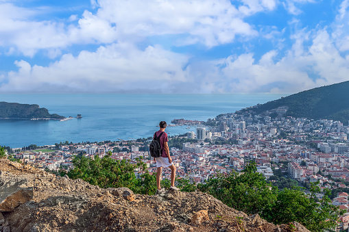 A male tourist with a backpack stands on top of a mountain and looks down at the city of Budva on the Adriatic Sea in Montenegro - rear view. A man stands on Viewpoint on Budva on the M2.3 highway