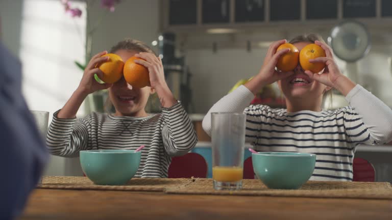 Family Having Fun at Breakfast: Parent and Daughters Making Funny Faces with Oranges. Playful Female Children Laughing While Having Essential Healthy Breakfast. Concept of Growing Up and Childhood