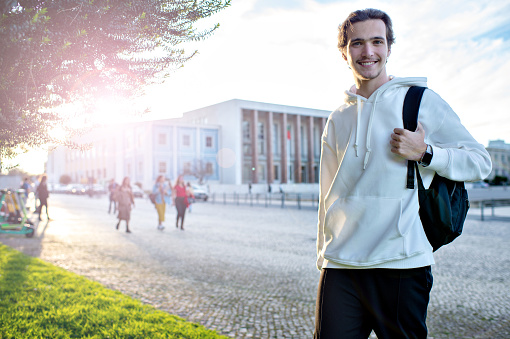 Student smiling while standing with a backpack on campus at his university. Happy man standing on a sidewalk near his faculty building. A cheerful young man with a backpack outdoors.