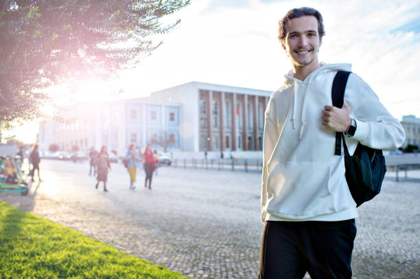 Student smiling while standing with a backpack on campus at his university. Happy man standing on a sidewalk near his faculty building. A cheerful young man with a backpack outdoors. stock photo