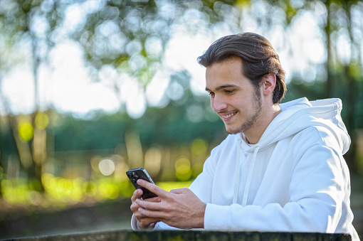 Young smiling guy looks at the screen of a cell phone while sitting in a park on a summer day. Portrait of an attractive young man with a phone in his hands outdoors. Happy white man with smartphone.