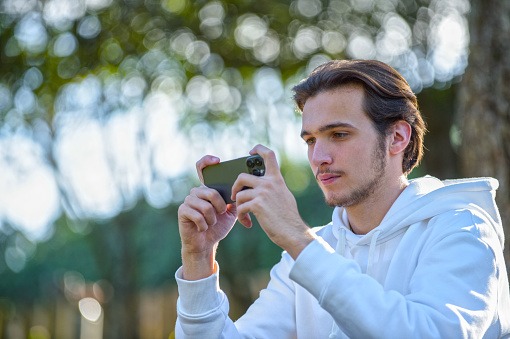 Focused young guy  with mobile phone in the park. Young man looking at cellphone screen outdoors. Serious caucasian  man looks at smartphone in his hand.