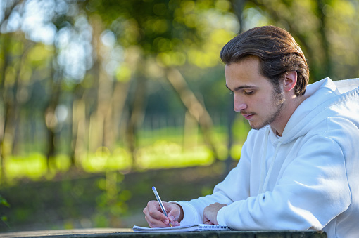 Student studies in nature. A young guy is studying outdoors sitting in the park. A young man writes with a pen in a notebook at a table in the park