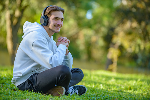 Portrait of young man in headphones listen to the music outdoors. Young smiling guy with headphones sitting on green lawn in the park and relaxing. Happy young man rests listening the music on nature