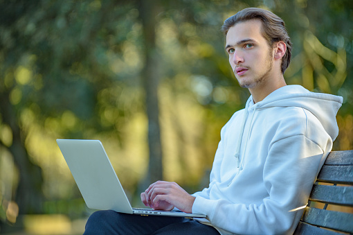 Portrait of thinking student at the park with laptop on his legs looks away.  Student with a computer in the park sitting on the bench, looking throu distance. Dreaming young man in nature, outdoors.