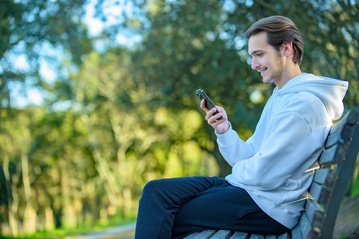 Young smiling guy looks at the screen of a cell phone while sitting on bench in a park. Portrait of an attractive young man with a phone in his hands outdoors. Happy white man with smartphone.