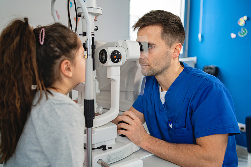 Caucasian male ophthalmologist examining with slit lamp structures of the eye, such as the cornea, iris, and lens, on a little girl at the ophthalmology clinic