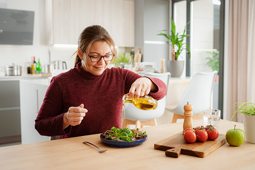 Portrait of a smiling woman dressing on healthy green salad with olive oil. She is sitting at dining table with modern kitchen at background. High resolution 42Mp indoors digital capture taken with SONY A7rII and Zeiss Batis 40mm F2.0 CF lens
