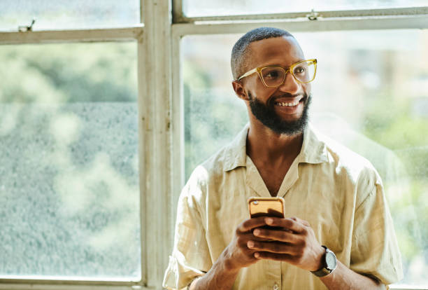 istock Image of a young African black man wearing glasses while holding a mobile phone and texting while texting and looking off camera with copy space. Stock Photo 1475573945
