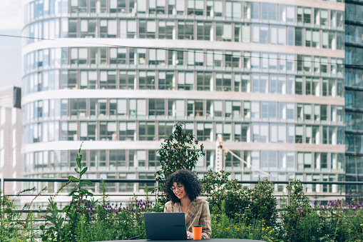 smiling african american woman sitting in front of the laptop on the terrace of her office in the banking district of the city
